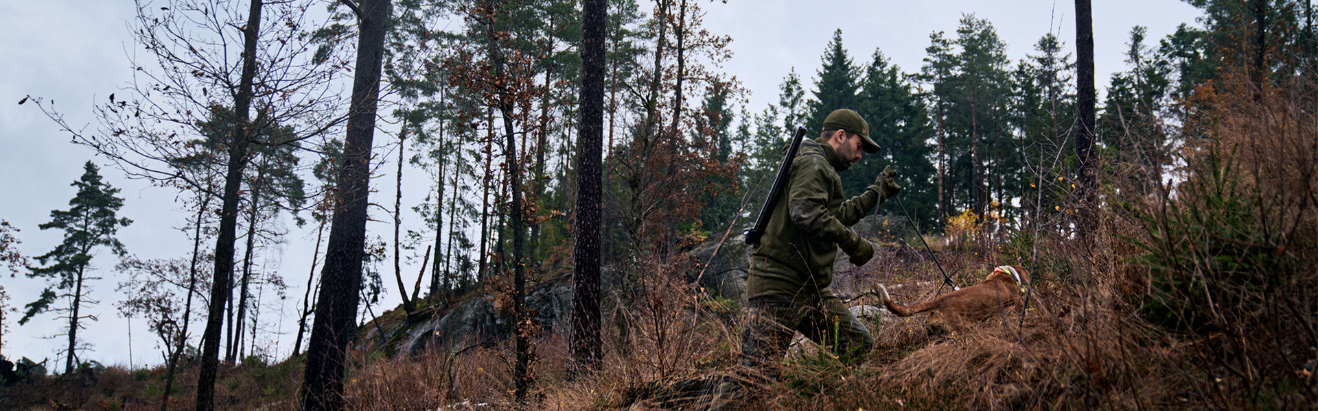 A man, in full hunting gear, walking uphill through some scrubby undergrowth